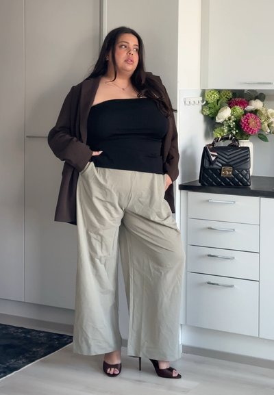 Mujer de pie en una cocina, vestida con un top negro sin tirantes, blazer marrón, pantalones anchos beige y tacones oscuros abiertos en los dedos.