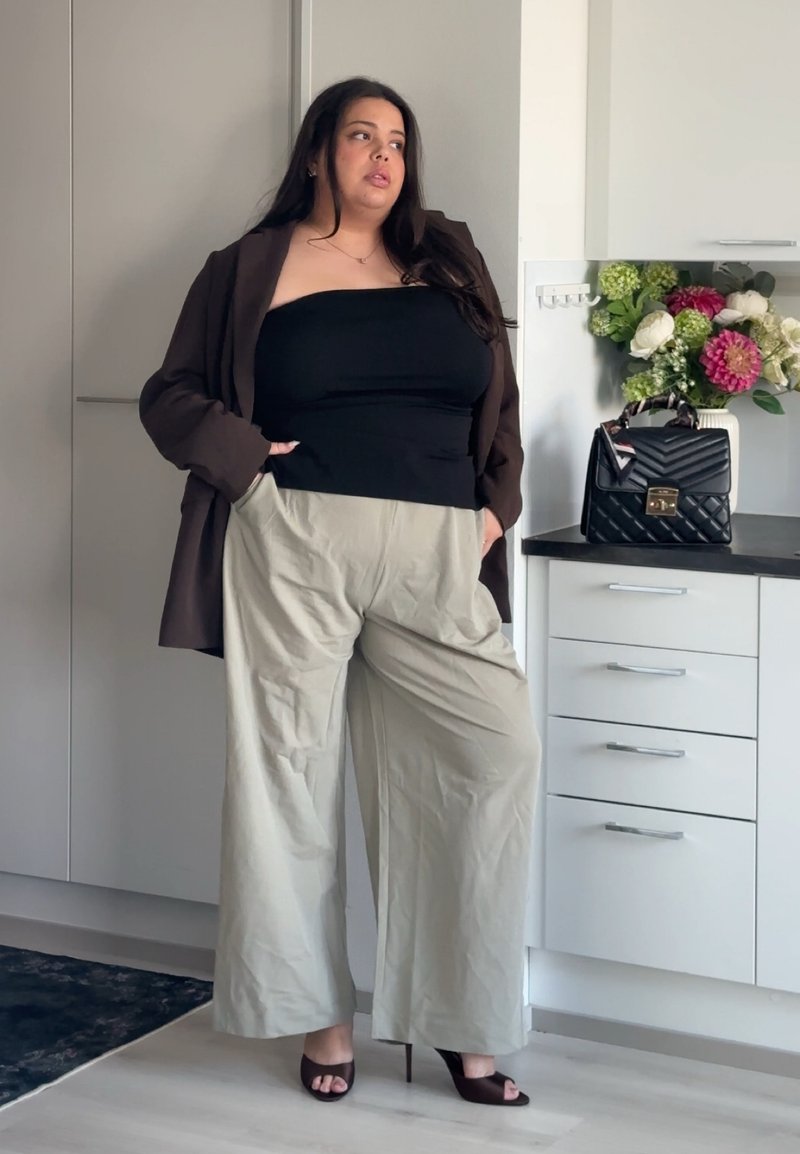 Femme debout dans une cuisine, portant un haut noir sans bretelles, un blazer marron, un pantalon large beige et des talons ouverts foncés.