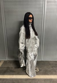 Sølvreflekterende oversized træningsdragt med lodrette striber, lange ærmer, høj krave og matchende taske. Baggrunden har et gittermønster.