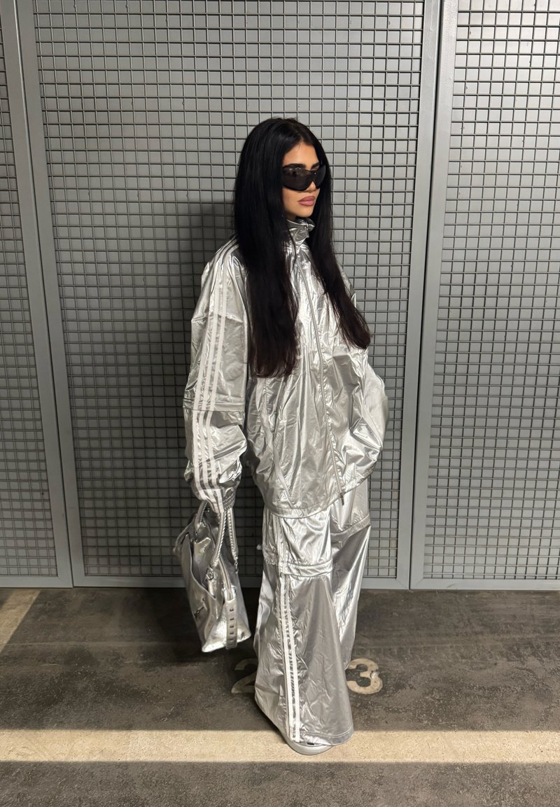 Silver reflective oversized tracksuit with vertical stripes, long sleeves, high collar, and matching bag. The background features a grid pattern.
