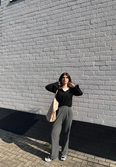 Mujer de pie al aire libre contra una pared de ladrillos blancos, vestida con una blusa negra, pantalones grises, zapatos blancos y sosteniendo un gran bolso beige con estampado.