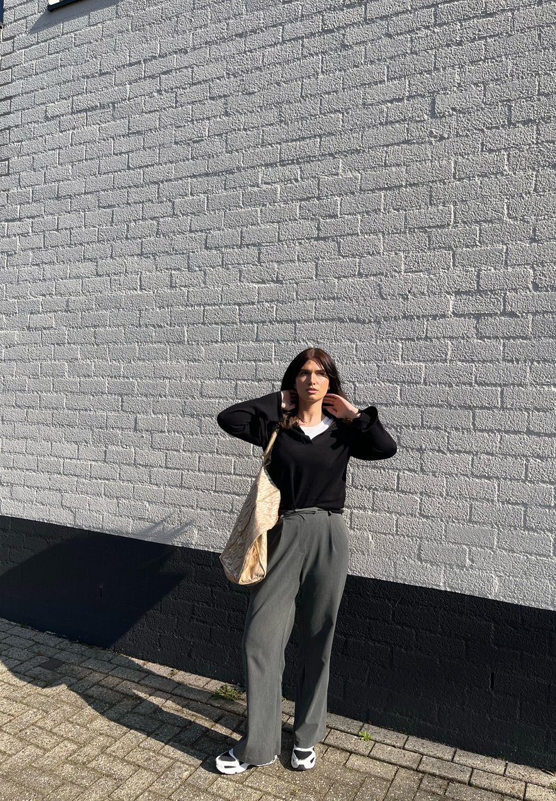 Frau steht im Freien vor einer weißen Backsteinwand, trägt ein schwarzes Oberteil, graue Hosen, weiße Schuhe und hält eine große beige Tasche mit Muster.