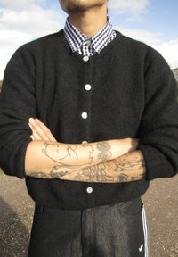 Černý vlněný kardigan s bílými knoflíky, vrstvený přes modrou a bílou kostkovanou košili. Paže jsou zkřížené a zobrazují různé tetování.