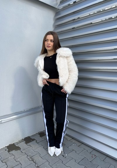 Chaqueta de piel sintética blanca con capucha, top corto negro, pantalones negros con rayas blancas y tacones blancos puntiagudos, contra una pared texturizada.