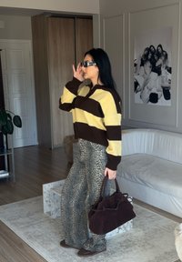 Pulôver de malha com riscas amarelas e castanhas, combinado com calças de padrão leopardo texturizado. Segurando uma mala castanha escura, usando óculos escuros escuros.