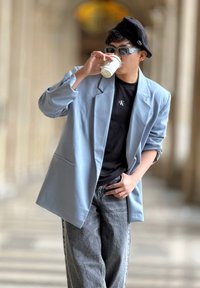 Heledu sinine suur lõikega bleiser, must T-särk logo jaotisega, hallid laiad teksad, must beret ja prillid. Hoides ühte ühekordset kohvikuppi.