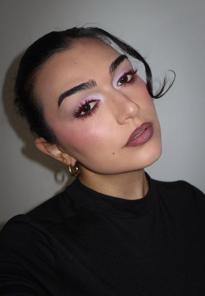 Retrato de una persona con cabello oscuro y piel clara, vestida con un suéter negro de cuello alto. El maquillaje presenta sombra de ojos rosa y un color de labios apagado. Pendientes de oro.