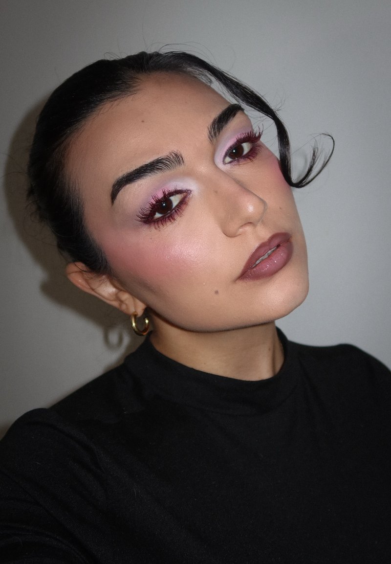 Ritratto di una persona con capelli scuri e pelle chiara, che indossa un dolcevita nero. Il trucco presenta ombretto rosa e un colore labbra tenue. Orecchini dorati.