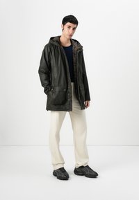 Groene waterdichte jas met een capuchon, voorzien van grote zakken aan de voorkant en een geruite voering. Draagt over een marineblauwe top en beige broek.