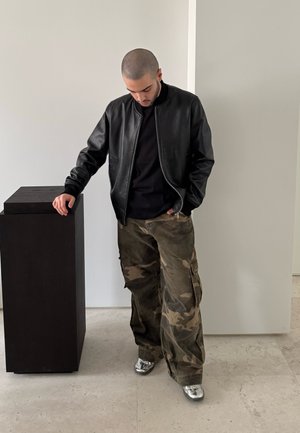 Chaqueta bomber de cuero negra sobre un suéter negro, pantalones cargo de camuflaje y zapatillas plateadas. De pie junto a un pedestal negro contra una pared clara.