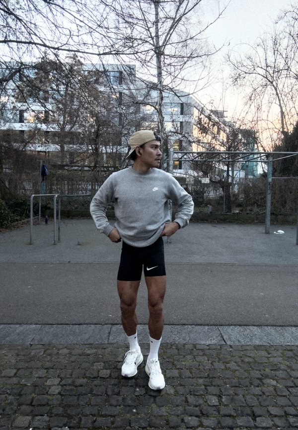 Vyras pilku džemperiu ir juodais šortais stovi ant akmenukais grįsto tako, rankos ant klubų, žvelgia į dešinę, už jo matosi miesto parkas ir pastatai.