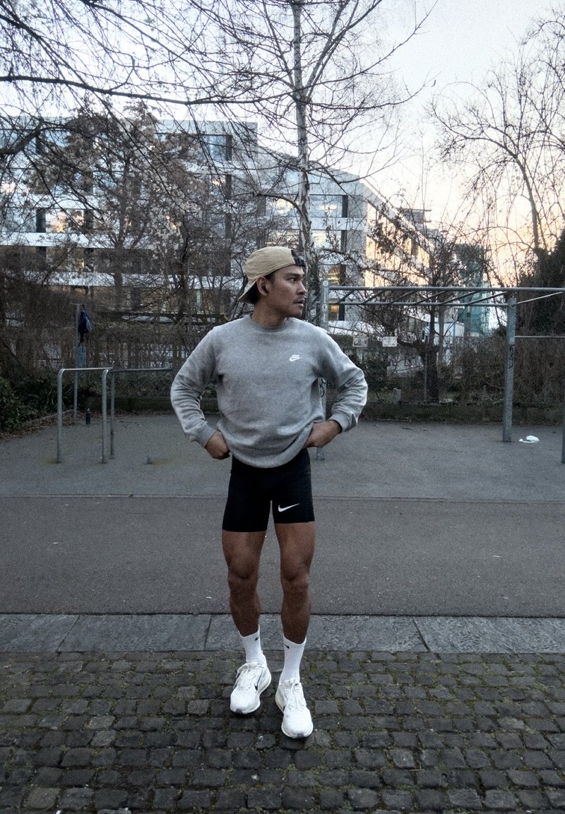 Ανδρας με γκρι φούτερ και μαύρο σορτς στέκεται σε πλακόστρωτο μονοπάτι, με τα χέρια στη μέση, κοιτάζει δεξιά, με αστικό πάρκο και κτίρια στο βάθος.