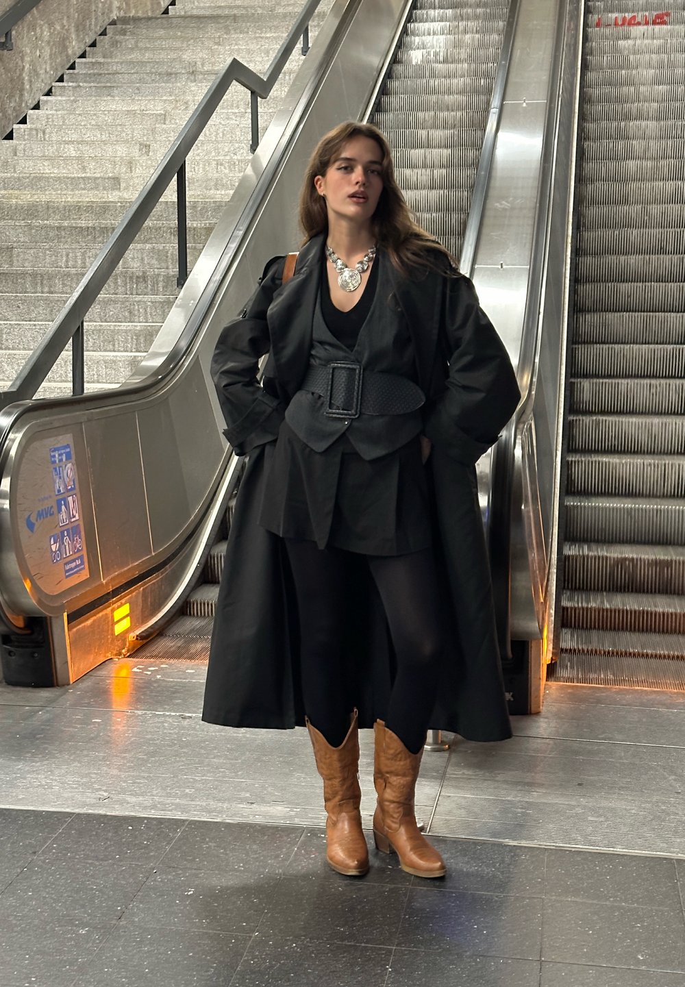 Palton negru supradimensionat cu mâneci largi peste o ținută neagră. Cizme cowboy maro. Colier din argint statement. Escalator în fundal.