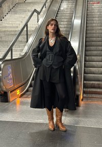 Palton negru supradimensionat cu mâneci largi peste o ținută neagră. Cizme cowboy maro. Colier din argint statement. Escalator în fundal.