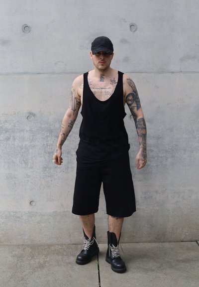 Top sin mangas negro y pantalones cortos, brazos tatuados, llevando una gorra negra y botas de combate negras con cordones blancos, de pie contra una pared de concreto.