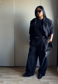 Blazer noir oversize sur une chemise noire, pantalon à jambes larges et petit sac à main. Les accessoires incluent des lunettes de soleil noires et une montre-bracelet.