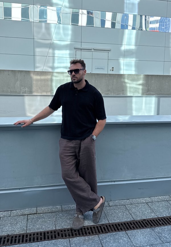 Férfi, aki egy szürke felületnek dől, fekete rövid ujjú pólót és barna vászonnadrágot visel. Napszemüvege és szürke lábbelije van.