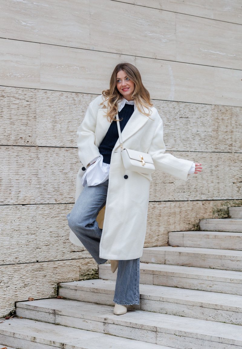 Cappotto in lana bianca con vestibilità morbida, bottoni neri, indossato sopra un maglione blu navy. Jeans a zampa di elefante azzurri e una borsa color crema completano il look.