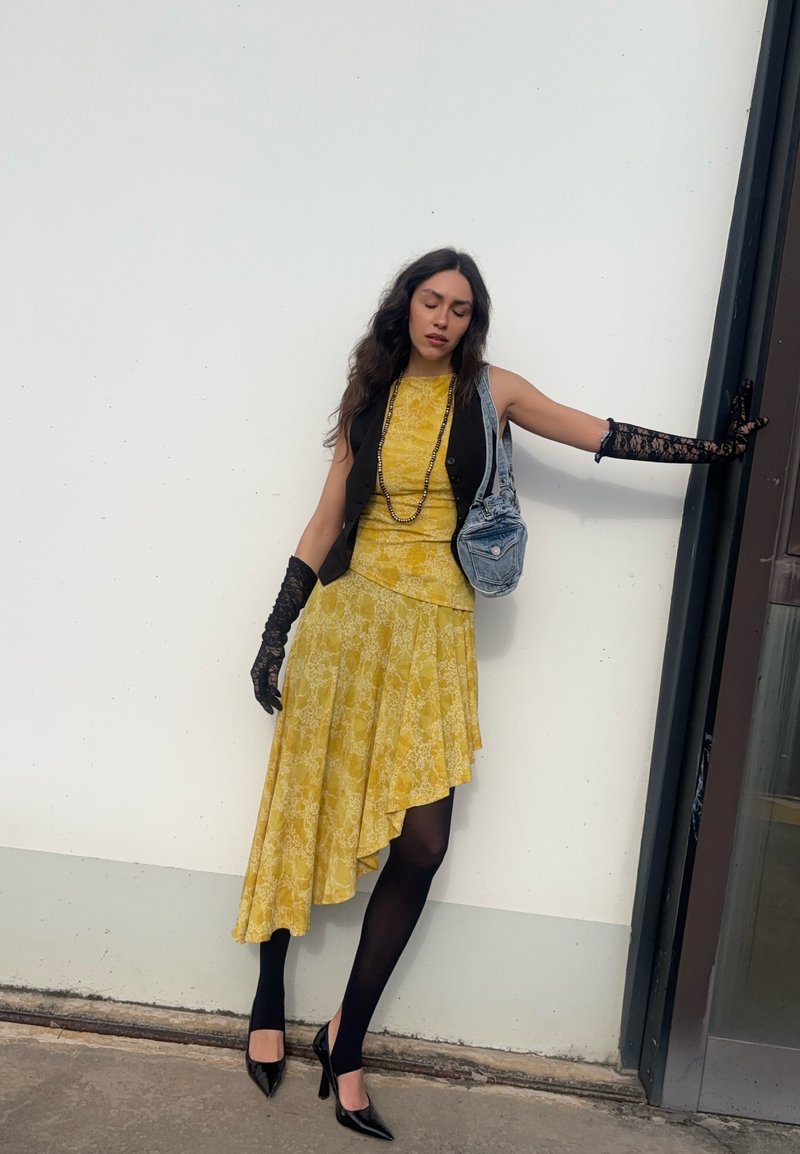 Femme en robe asymétrique jaune, gilet noir, gants en dentelle, collants noirs et talons appuyée contre un mur blanc près d'une porte.