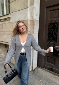 Sivý nadrozmerný kardigán s výstrihom do V, štruktúrovaná tkanina, široké rukávy. Kombinovaný s bielym topom a džínsami s vysokým pásom, drží šálku kávy.