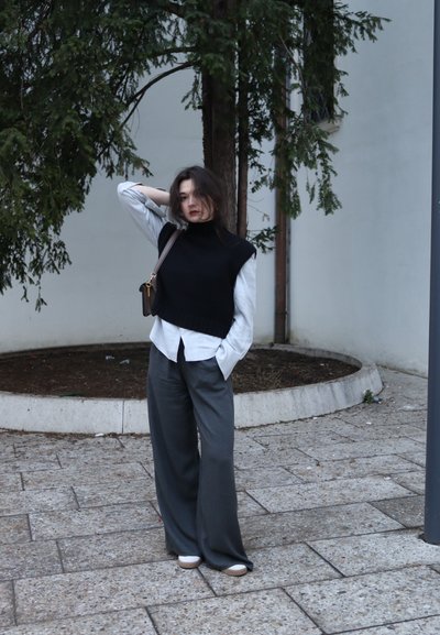 Suéter negro sin mangas sobre una camisa abotonada gris claro, combinado con pantalones anchos grises y zapatos planos beige. Entorno al aire libre con vegetación.