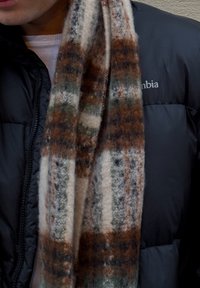 Cachecol de lã com padrão xadrez em bege, verde e ferrugem, colocado sobre uma jaqueta puffer preta com costuras visíveis e um fecho de correr.