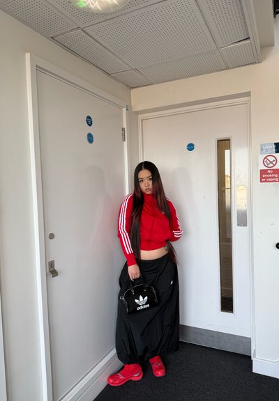 Chaqueta roja con cremallera y rayas blancas, pantalones holgados negros, zapatos rojos y un bolso negro con un logo blanco. Paredes blancas y suelo gris visibles.