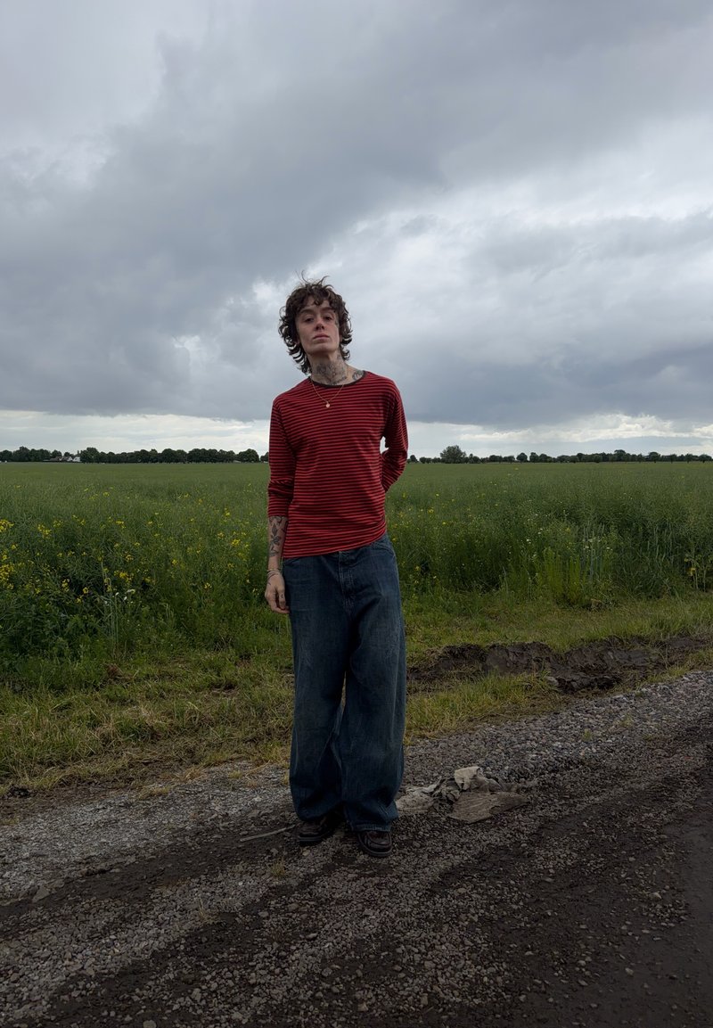 Jeune personne avec des tatouages portant un t-shirt rayé rouge et un jean ample, debout sur une route en gravier à côté d’un champ vert sous un ciel nuageux.