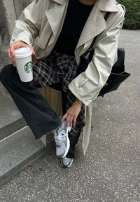 Beige trenčkot přes černé tričko, kalhoty s kostkovaným vzorem, černé džíny a šedé tenisky. Drží bílý kelímek Starbucks.