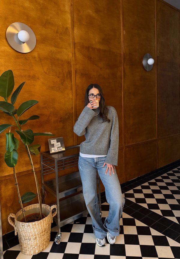 Kvinde med briller, grå sweater og jeans, der drikker af en kop, stående ved siden af en plante og en metalvogn på sort-hvidt flisegulv.