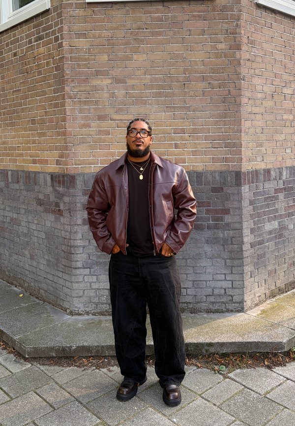 Man with glasses and braided hair wearing a brown leather jacket, black shirt, loose black pants, and boots standing by a brick corner wall.