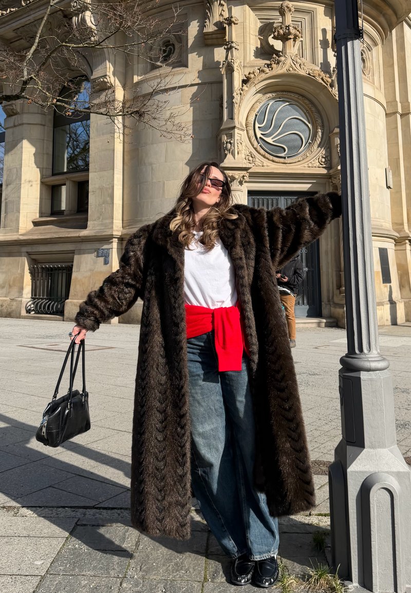 Femme portant un long manteau en fourrure sombre, chemise blanche, pull rouge noué autour de la taille, jean bleu, chaussures noires, lunettes de soleil, tenant un sac à main noir près d’un bâtiment orné.