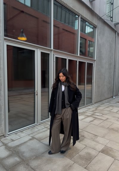 Abrigo largo negro, bufanda gris, chaleco negro y pantalones de pierna ancha beige. De pie sobre pavimento de piedra con grandes ventanales al fondo.