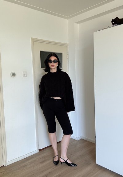 Mujer con suéter negro, pantalones cortos hasta la rodilla, tacones puntiagudos y gafas de sol de ojo de gato, de pie en el interior sobre suelo de madera.