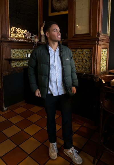 Chaqueta de plumón verde, camisa de rayas azul claro, pantalones negros y zapatillas beige. Presenta una tela texturizada y una silueta ajustada.