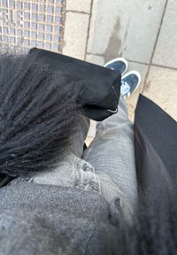 Personne portant un jean gris, un manteau noir et des chaussures noires, debout sur un trottoir carrelé, tenant un sac noir texturé.