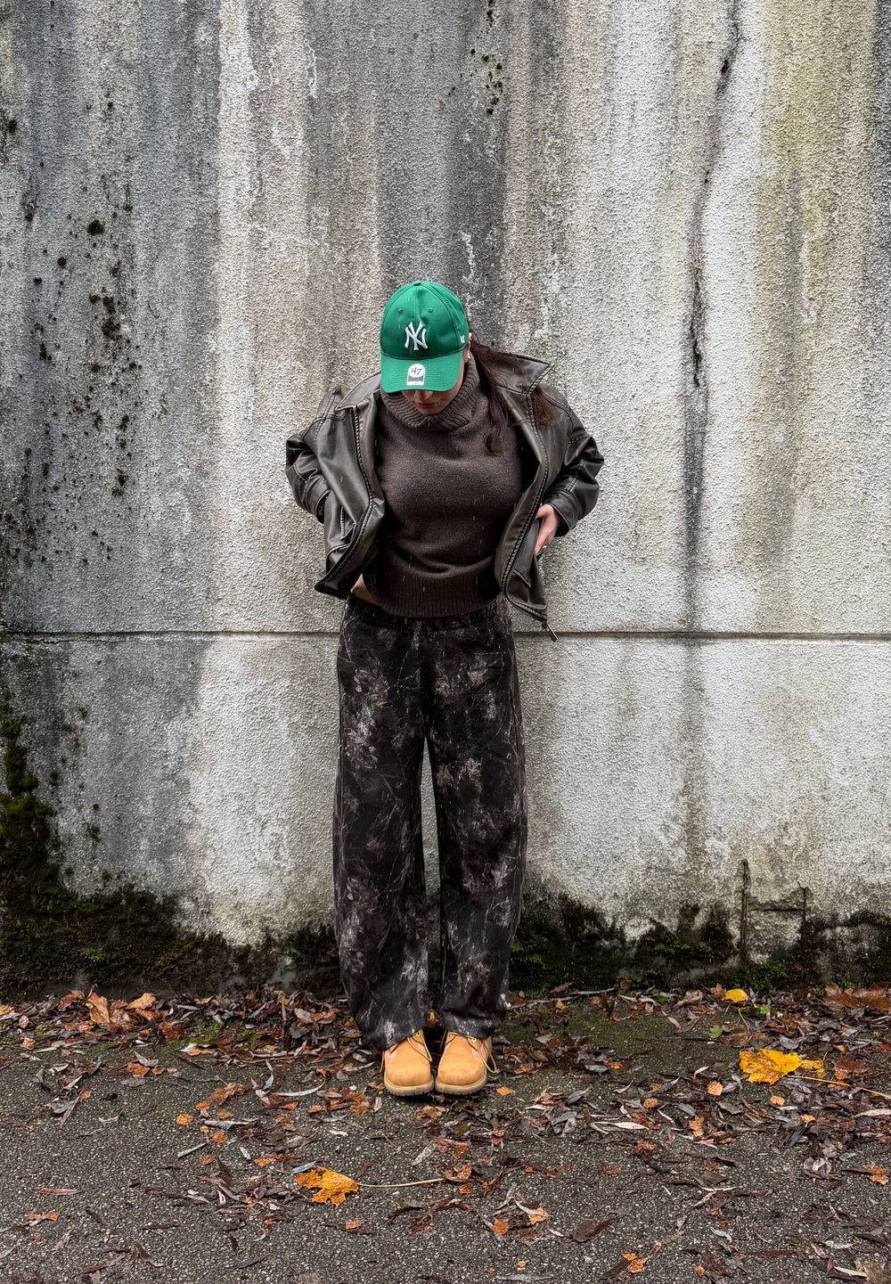 Osoba v zelenej čiapke, hnedej koženej bunde, tmavom svetri, vzorovaných nohaviciach a béžových topánkach stojaca pri ošúchanom betónovom múre s listami na zemi.