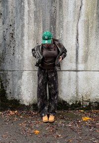 Osoba v zelenej čiapke, hnedej koženej bunde, tmavom svetri, vzorovaných nohaviciach a béžových topánkach stojaca pri ošúchanom betónovom múre s listami na zemi.