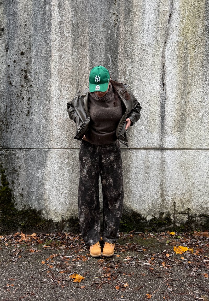 Pessoa a usar um boné verde, casaco de cabedal castanho, camisola escura, calças com padrão e botas cor de castanho claro, em pé junto a uma parede de betão desgastada, com folhas no chão.