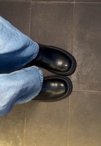 Άτομο που φοράει χαλαρά ανοιχτό μπλε τζιν και μαύρα δερμάτινα μπότες, στέκεται σε σκοτεινό πλακάκι δάπεδο.