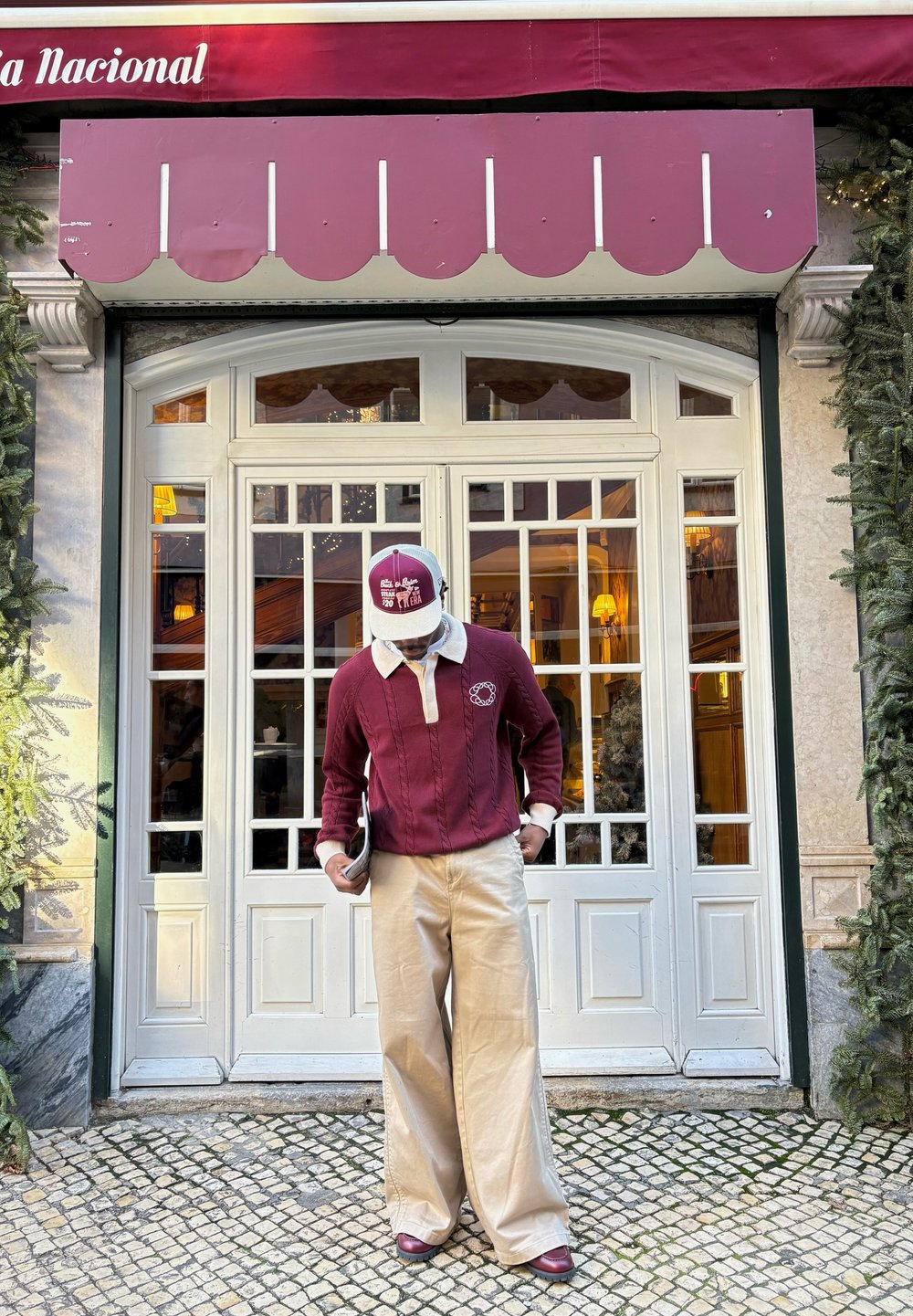 Homme en pull bordeaux et casquette portant un pantalon large beige, debout dans une rue pavée devant des portes-fenêtres blanches sous un auvent bordeaux.
