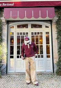 Homme en pull bordeaux et casquette portant un pantalon large beige, debout dans une rue pavée devant des portes-fenêtres blanches sous un auvent bordeaux.