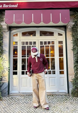 Hombre con suéter burdeos y gorra, pantalones anchos beige, de pie en una calle empedrada frente a puertas francesas blancas bajo un toldo burdeos.