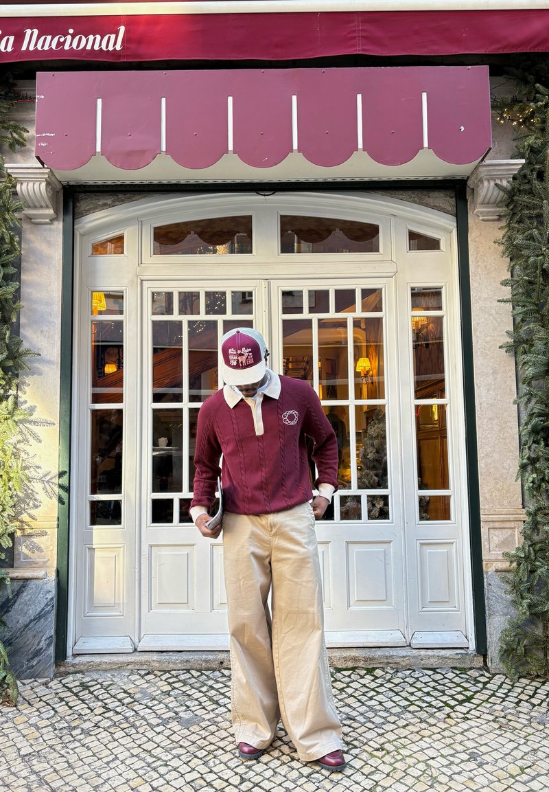 Homme en pull bordeaux et casquette portant un pantalon large beige, debout dans une rue pavée devant des portes-fenêtres blanches sous un auvent bordeaux.