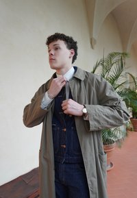 Χακί καμπαρντίνα πάνω από ένα σκούρο τζιν ολόσωμο, λευκό πουκάμισο και πράσινη ριγέ γραβάτα. Τα αξεσουάρ περιλαμβάνουν ένα ασημί ρολόι και έναν απλό φόντο.