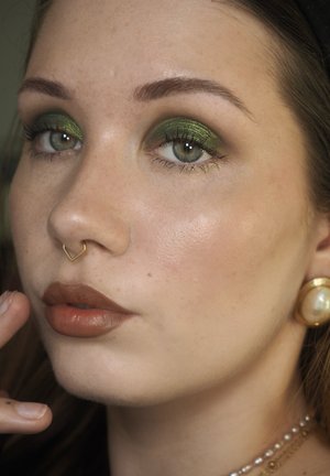 Close-up of a face featuring green shimmer eyeshadow, a bronze lip color, a gold heart-shaped nose ring, and a large pearl earring.