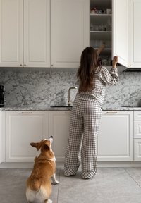 Femme en pyjama à carreaux atteignant un placard de cuisine en hauteur tandis qu'un chien brun et blanc observe depuis le sol.