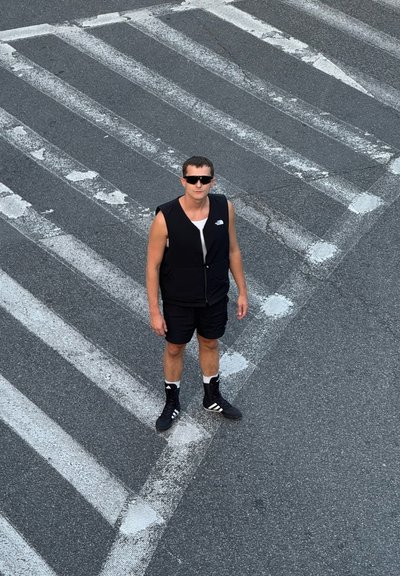 Chaleco acolchado negro sobre una camiseta blanca, pantalones cortos negros y zapatillas altas negras. De pie en un paso de peatones gris con franjas blancas.