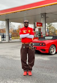 Červené polo s logom Ferrari, hnede voľné nohavice, červené tenisky a slnečné okuliare. Doplnky zahŕňajú opasok a prívesky. V pozadí je čerpacia stanica.