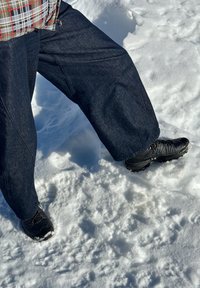 Personne portant un jean foncé et des bottes noires marchant dans la neige, une jambe levée, habillée pour le temps froid.